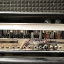 Fotografie zachycuje vnitřek kytarového komba s otevřeným chassis, kde jsou vidět elektronické lampy a zapojení. Na chromovaném předním panelu je náp…