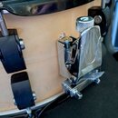 Detailní pohled na dřevěný korpus bubnu světlé barvy s černým obručem. Na korpusu jsou připevněny černé kovové držáky a chromovaný mechanismus s knur…