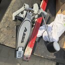 Šedý kovový pedál basového bubnu s černým plastovým krytem, na kterém je nápis ALESIS. Pedál má kovovou tyč s černou gumovou koncovkou a je položen n…