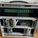 Modulární syntetizátor v hliníkovém kufru s černou rukojetí. Na horním a dolním rámu je nápis A-100 ANALOG MODULAR SYSTEM a logo DOEPFER. V systému j…