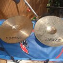 Dvě činely z mosazi položená na modré transportní tašce s červeným zipem a logem "F". Levý činel nese nápis "istanbul Agop Handmade cymbals from Turk…