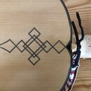 Detail dřevěného těla strunového nástroje světlé barvy s viditnou sletovkou. Na povrchu je vyrytý nebo vykreslený geometrický ornament tvořený prople…