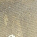 Detailní záběr na povrch hudebního nástroje z matného, hnědlého dřeva s viditelnou strukturou jemných vodorovných rýh. Na povrchu je vyryt nápis „Tim…
