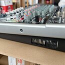 Stříbro-černý mixážní pult s řadou potenciometrů v různých barvách (šedá, oranžová, červená) a konektory na pravé straně. Na horním panelu je vidět l…