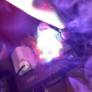 Fotografie zachycuje elektronické zařízení s černým šasi, které vyzařuje intenzivní bílé a barevné světlo doprovázené hustým kouřem. Na horní části j…