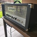 Černý kytarový kombozesilovač s kovovou mřížkou a stříbrným ovládacím panelem. Na mřížce je nápis LAMP a na spodním panelu text GUITAR TUBE AMPLIFIER…