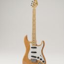 Elektrická kytara s přírodním dřevěným povrchem těla a hmatníkem. Na hlavě je viditelné logo "Suhr". Nástroj má bílý pickguard, tři snímače typu sing…