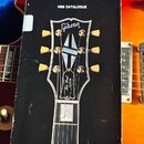Fotografie zachycuje černý katalog s nápisem 1988 CATALOGUE, který leží na elektrické kytáře v modrém pouzdře. Na obálce katalogu je zobrazen hриф hl…