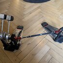 Na fotografii je dvojpedál k basové bubnu v kovovém provedení s černými a červenými prvky. Na pedálech jsou viditelné nápisy "Trickfoot" a "DW". Kons…