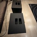 Dvě černé bedny s texturovaným povrchem, pravděpodobně reproduktory nebo subwoofery. Každá bedna má na horní straně dva obdélníkové otvory. Na fotogr…