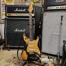 Elektrická kytara typu Stratocaster s výrazně opotřebovaným černým lakem, pod kterým je vidět žlutý povrch. Kytara má světlý javorový hmatník, tři sn…