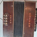 Hnědý akordeon s dřevěným povrchem a černým měchu. Na pravé straně je svisle napsán nápis HOHNER a na levé straně nápis ERIKA, oba v oranžové barvě.…