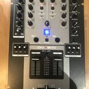 Šedý DJ mixér s černým šasi a nápisem DENOM v horní části. Na přístroji jsou vidět ovládací prvky, modře podsvícené tlačítko a v dolní části modelové…