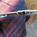 Detailní pohled na horní část stříbrné flétny z kovu. Na trubici je vidět malý trojúhelníkový symbol a kulaté logo s hvězdicí. Povrch je lesklý s vid…