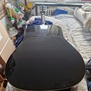 Zadní strana akustické kytary v černé barvě s vysokým leskem. Povrch vykazuje drobné škrábance a prachové částice. Na fotografii není vidět žádný náp…