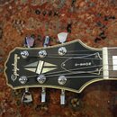 Fotografie zachycuje hlavu elektrické kytary v černé barvě s bílým lemováním. Na hlavě je zleva doprava nápis Epiphone, stylizovaný kosočtvercový sym…