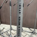 Detail šedého kovového stojanu s nápisem kÖNIG & MEYER »STARLINE«. Nástroj má teleskopické nohy a je umístěn na dlažbové ploše před hnědými dveřmi. P…