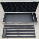 Otevřený hliníkový transportní kufr s černou vnitřní výplní a vátovou pěnou v horní části víka. Před kufrem leží černý, mřížkovitý vložný element s m…