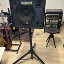 Černý kombozesilovač značky Roland s nápisem KC na spodní části mřížky. Zařízení je umístěno na černém kovovém stativu s trojnožkou. V pozadí jsou vi…