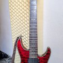 Červená elektrická kytara s výraznou dřevěnou strukturou povrchu a černým lemováním. Na hlavě hmatníku je bílý nápis "Schecter". Kytara má dva humbuc…