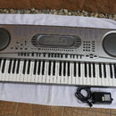 Stříbrošedý digitální klavír značky CASIO s nápisem MUSICAL INFORMATION SYSTEM. Nástroj má v horní části displej, řadu ovládacích tlačítek a dva velk…