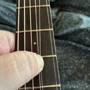 Detailní pohled na hриф kytary s tmavým dřevěným hmatníkem a kovovými pražci. Na hmatníku jsou vidět bílé tečkové značky poloh. Prst ruky přitlačuje…