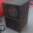 Dvě černé, kubické basové reproduktory v texturovaném povrchu, které jsou naskládány na sebe. Přední strany jsou kryty kovovými perforovanými mřížkam…