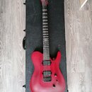 Červená elektrická kytara s matným povrchem položená na černém tvrdém kufru. Na hlavě hmatníku je bílý nápis "Schecter". Nástroj má dva humbuckery, j…