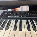 Detailní pohled na klávesový nástroj s černým povrchem a bílo-černými klávesami. Na ovládacím panelu je nápis Organ/Strings O9 a RS-09. Panel obsahuj…