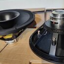 Dvě černé reproduktorové membrány s kovovými magnety, položené na kartonových krabicích. Levý reproduktor má uprostřed vyvýšený kuželovitý středový h…
