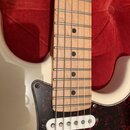 Detail elektrické kytary s bílým trupem a hnedý hmatníkem z jasného dřeva s černými tečkovými značkami. Kytara má tmavě červený pickguard s texturou…