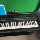 Černý digitální syntezátor v černém kufru s zeleným vnitřním polstrováním. Na horním panelu je vidět nápis DX7 a tabulky s parametry. V kufru vedle n…