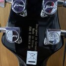 Záběr na zadní stranu černé hlavy kytary s chromovanými ladicími kolíky. Na každém z kolíků je vyryto logo Framus. Uprostřed hlavy je svislý nápis De…