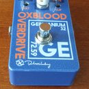 Modrý efekt pedál s nápisy OXBLOOD a OVERDRIVE. Na bílém obdélníkovém poli je uveden text GERANIUM 32, GE a číslo 72.59. Ve spodní části je bílý podp…