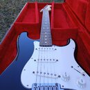 Tmavě modrá elektrická kytara typu Stratocaster s bílým pickguardem a třemi snímači. Nástroj leží v otevřeném kufru s červeným polstrováním. Na ovlád…