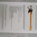 Fotografie zobrazuje pětistrunnou elektrickou basovou kytaru v bílé barvě s hnědým hmatníkem. Na hlavě nástroje je černý nápis Fender a modelové ozna…