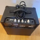 Černý basový kombozesilovač s texturovaným povrchem a přiloženým napájecím kabelem. Na zadním panelu je nápis CM20B 20W BASS AMPLIFIER, značka Cort a…