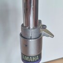 Detail chromovaného kovového stojanu s regulačním mechanismem. Na šedém plastovém prstenci je fialový nápis YAMAHA a pod ním menší bílý text MADE IN…