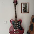 Červená elektrická kytara s polodupatým korpusem a f-otvory, zavěšená na bílé stěně. Nástroj má tmavý hmatník, černý pickguard a stříbrný vibrační sy…