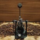 Fotografie zobrazuje černý basový pedál s kovovými mechanismy a řetězovým pohonem. Na horní části je umístěn kulatý gumový stomeč. Zařízení stojí na…