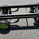 Černý transportní vozík s kovovou konstrukcí a zelenými detaily. Má dvě velká pneumatická kola vpředu a dvě menší otočná kolečka vzadu. Na zelených p…