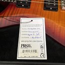 Detailní záběr na tělo kytary s připevněným bílým papírovým štítkem. Na štítku je nápis PRS S2 Series Instruments are proudly made in the USA at the…