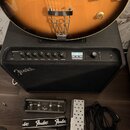 Fotografie zachycuje spodní část kytary, černý kytarový kombozesilovač Fender Mustang GTX a příslušenství. Na předním panelu zesilovače je nápis MUST…