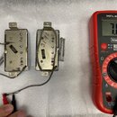 Dvě kovové součástky s vnitřními cívkami a připojenými vodiči, vedle nich červený digitální multimetr značky POWERFIX Profi s displejem ukazujícím ho…
