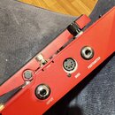 Detailní pohled na červený kovový panel hudebního zařízení. Na panelu jsou umístěny tři konektory s nápisy INPUT, MIDI a FOOTSWITCH. V horní části je…