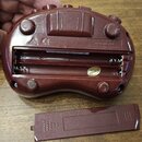 Hnědý plastový přístroj s otevřeným bateriovým slotem a vedle položeným víčkem. Na spodní části je vyražený text DESIGNED IN U.S.A., MADE IN CHINA a…
