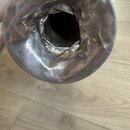 Detailní pohled do vnitřku kovového zvonu hudebního nástroje stříbrné barvy. Povrch kovu je v oblasti přechodu do trubice výrazně zdeformovaný, pomač…