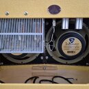 Detail vnitřku kytarového komba s dvěma reproduktory a bílou kovovou mřížkou zakrývající elektroniky. Na pravém reproduktoru je štítek s nápisy „Joe…