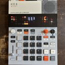 Bílo-šedý sampler s černým displejem a oranžovými detaily. Na horním bílém panelu je nápis K.O.II, pod ním japonské znaky サンプラー a text 64 MB SAMPLER…