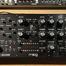Černý analogový syntetizátor v dřevěném rámu s nápisy LABYRINTH a PARALLEL GENERATIVE ANALOG SYNTHESIZER. Uprostřed spodní části je bílé logo moog. P…
